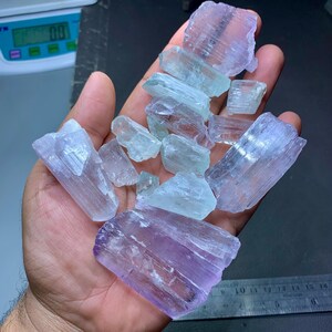 May include: A hand holding a collection of raw, uncut gemstones. The stones vary in color, with shades of light purple, blue, and clear. The crystals have a rough, natural texture and are irregularly shaped.