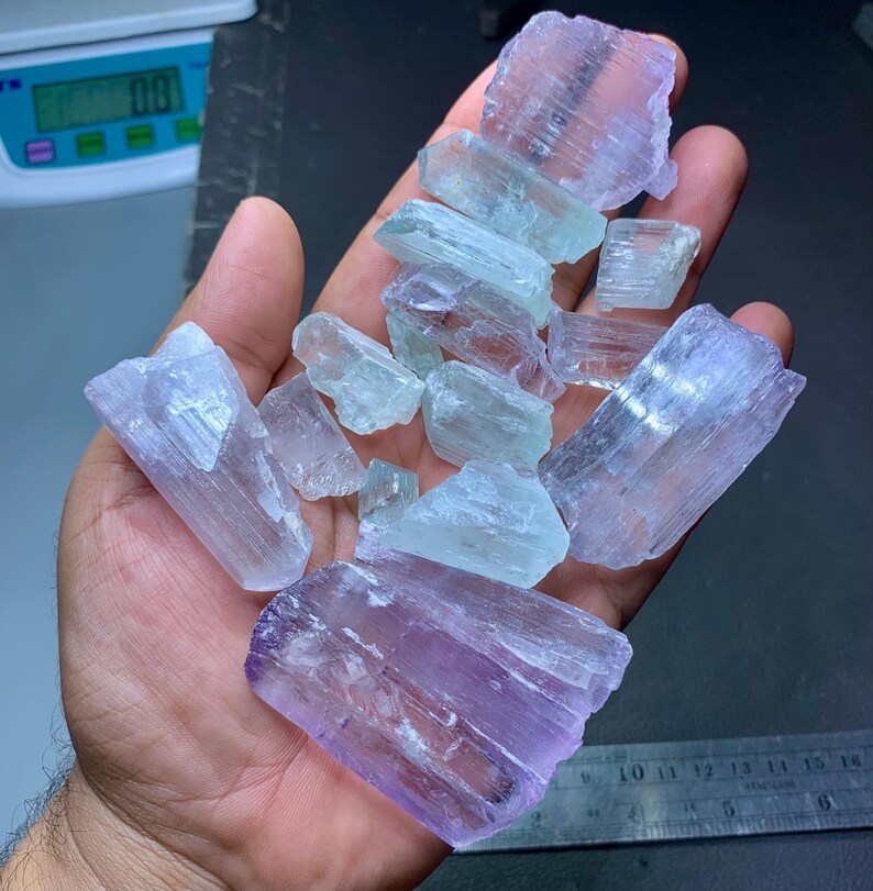 May include: A hand holding a collection of raw, uncut gemstones. The stones vary in color, with shades of light purple, blue, and clear. The crystals have a rough, natural texture and are irregularly shaped.