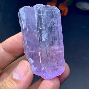 May include: A raw, natural crystal specimen in shades of lavender and purple. The crystal has a rectangular shape with visible striations and a slightly rough texture. The crystal is held in a hand, showcasing its size and color.