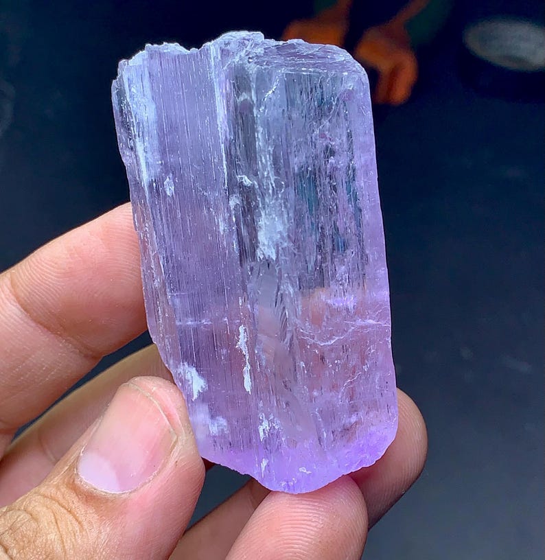 May include: A raw, natural crystal specimen in shades of lavender and purple. The crystal has a rectangular shape with visible striations and a slightly rough texture. The crystal is held in a hand, showcasing its size and color.