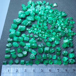 Op de afbeelding: Een verzameling ruwe, ongeslepen smaragden in verschillende maten, verspreid over een grijs oppervlak. De edelstenen vertonen een levendige groene kleur en zijn in de buurt van een liniaal geplaatst voor de schaal. De liniaal toont metingen in centimeters.