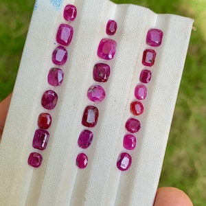 May include: Rows of faceted, deep red rubies of varying shapes and sizes are displayed on a white surface. The gemstones range from oval to square cuts, showcasing their rich color and clarity. The arrangement highlights the natural beauty of the precious stones.