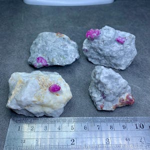May include: Four rough-cut gemstones with vibrant pink ruby inclusions. The stones are set against a gray surface, with a ruler providing scale. The gemstones are a mix of gray and white, with the ruby crystals adding a pop of color.