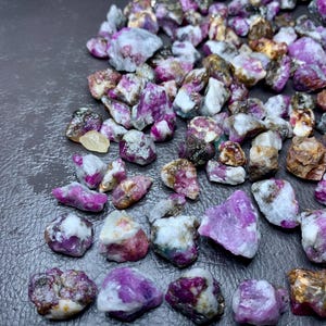 May include: A collection of raw ruby gemstones in various sizes and shapes. The stones display a range of colors, including deep pink, purple, and white, with some having a translucent appearance. A ruler is placed at the bottom, with markings in inches.