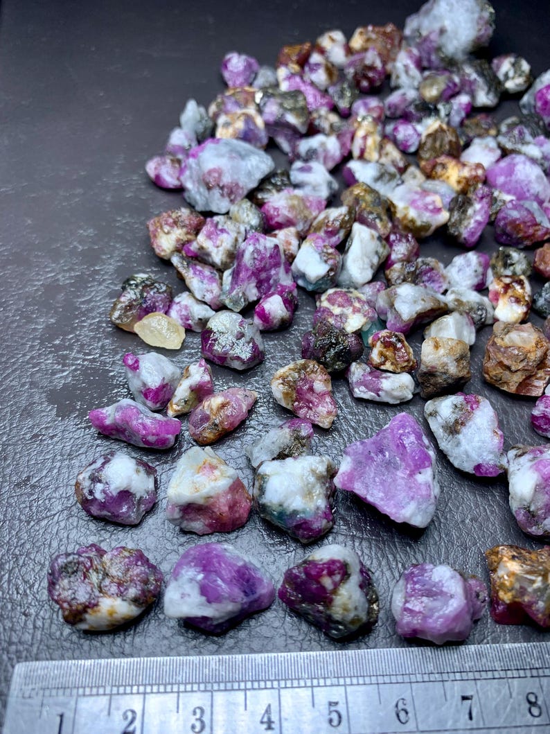 May include: A collection of raw ruby gemstones in various sizes and shapes. The stones display a range of colors, including deep pink, purple, and white, with some having a translucent appearance. A ruler is placed at the bottom, with markings in inches.