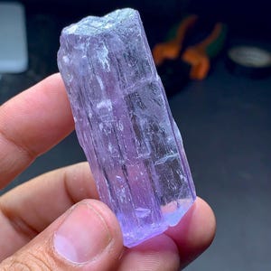 May include: A hand holds a raw, uncut, lavender-colored crystal. The crystal is rectangular with a rough, textured surface and a slightly translucent appearance. The crystal's color varies from pale to deep purple.