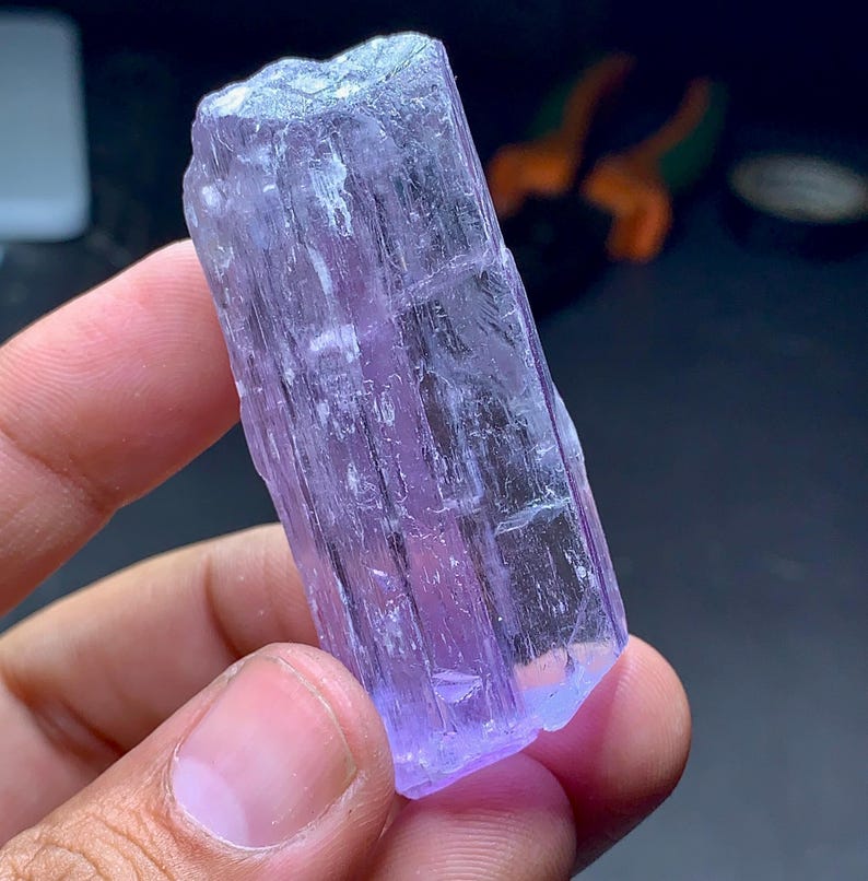 May include: A hand holds a raw, uncut, lavender-colored crystal. The crystal is rectangular with a rough, textured surface and a slightly translucent appearance. The crystal's color varies from pale to deep purple.