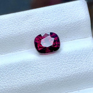 May include: A cushion-cut, faceted gemstone in shades of deep red and magenta. The gem is set against a white, textured background, showcasing its brilliance and clarity. The gemstone's facets reflect light, creating a sparkling effect.