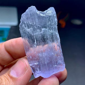 May include: A translucent, light purple crystal with a rough, textured surface. The crystal is held in a hand, showcasing its natural, irregular shape and subtle color variations. The crystal's surface has vertical striations.