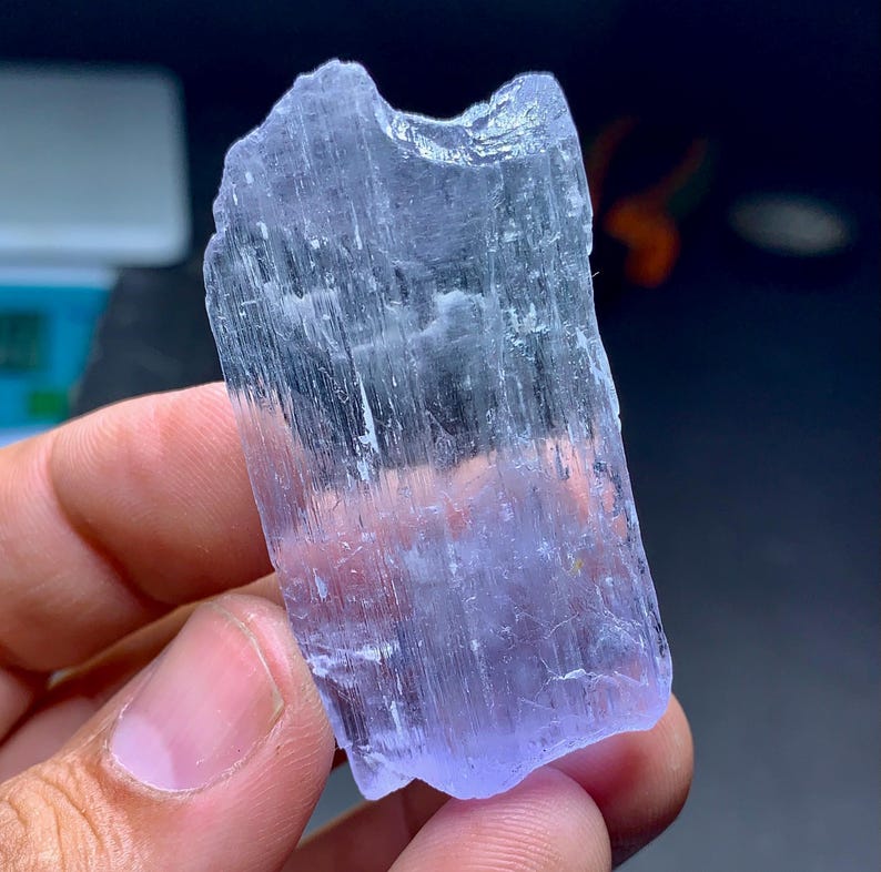May include: A translucent, light purple crystal with a rough, textured surface. The crystal is held in a hand, showcasing its natural, irregular shape and subtle color variations. The crystal's surface has vertical striations.