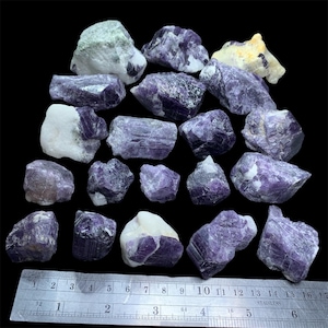 Peut inclure: Plusieurs cristaux d'améthyste bruts, de différentes tailles et formes, avec des nuances de violet et de blanc. Une règle en acier inoxydable est placée en bas, indiquant les mesures en centimètres. Les cristaux sont sur fond noir.