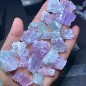 以下が含まれることがあります： 淡い青、緑、紫の色合いの、生の、未加工の宝石の束。結晶はざらざらとした自然な質感で、さまざまな形をしています。宝石は、暗い背景に対して開いた手の中に保持されています。