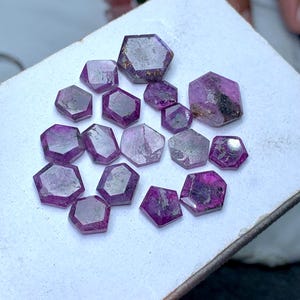May include: A collection of fifteen loose, faceted gemstones in varying shades of magenta and deep pink. Each stone features a distinct hexagonal or pentagonal shape with visible surface inclusions and a semi-translucent, polished finish.