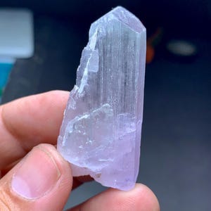 May include: A light purple crystal with a faceted top and a rough, textured surface. The crystal is held in a hand, showcasing its natural form and color variations. The crystal's color ranges from pale lavender to a translucent, almost clear hue.