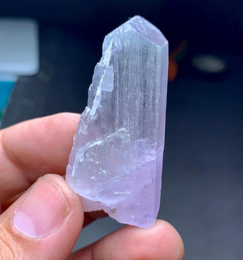 May include: A light purple crystal with a faceted top and a rough, textured surface. The crystal is held in a hand, showcasing its natural form and color variations. The crystal's color ranges from pale lavender to a translucent, almost clear hue.
