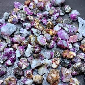 May include: A collection of raw ruby gemstones in various sizes and shapes. The stones display a range of colors, including deep pink, magenta, and white, with some having a translucent appearance. The rough texture of the stones is visible.
