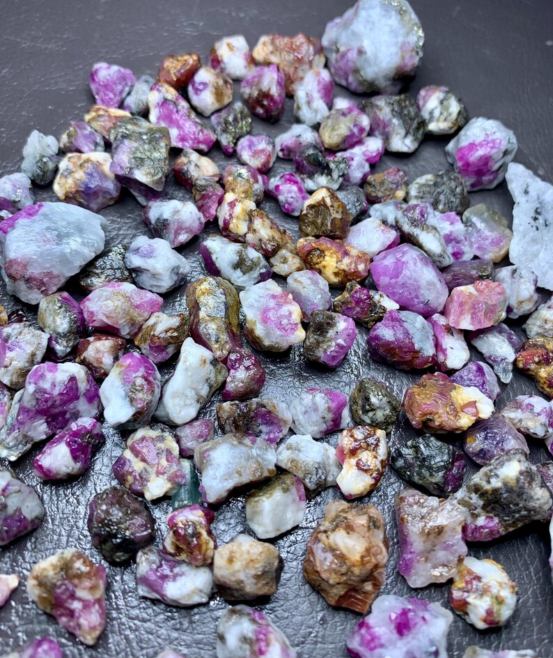 May include: A collection of raw ruby gemstones in various sizes and shapes. The stones display a range of colors, including deep pink, magenta, and white, with some having a translucent appearance. The rough texture of the stones is visible.