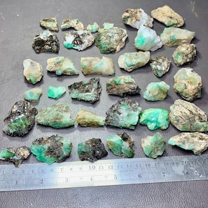 May include: A collection of rough emerald crystals with black inclusions. The crystals are various sizes and shapes, and some are partially covered in a clear, white matrix. The crystals are arranged on a gray surface with a ruler for scale.