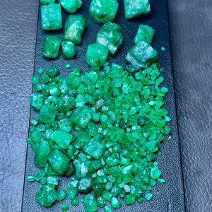 Op de afbeelding: Een verzameling ruwe smaragden, in verschillende maten en vormen, gepresenteerd op een zwart oppervlak. De smaragden hebben een levendige groene kleur, waarbij sommige meer doorschijnend lijken. De opstelling omvat grotere, ongesneden stenen en kleinere, gefragmenteerde stukken.