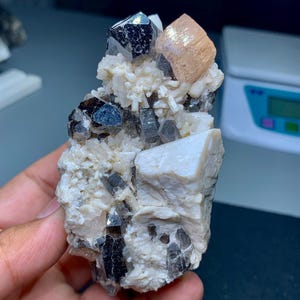 May include: A cluster of natural crystals featuring a variety of colors and textures. The focal point is a large, dark, lustrous crystal, complemented by smaller, smoky-colored crystals and a peach-colored crystal. The crystals are set in a matrix of white and beige minerals.