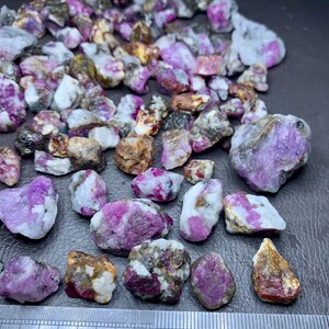 May include: A collection of raw ruby gemstones, displaying a mix of pink, purple, and white hues. The stones vary in size and shape, with a stainless steel ruler providing scale. The gemstones are rough-cut and natural.