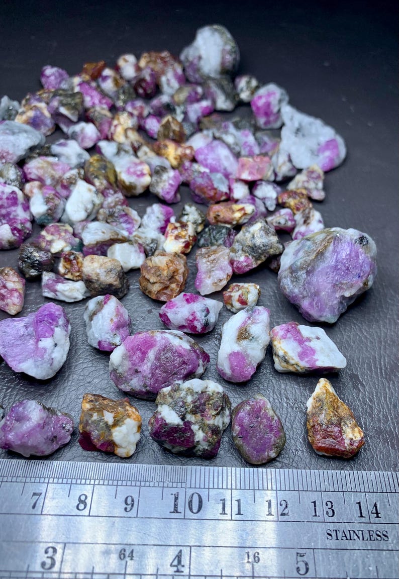 May include: A collection of raw ruby gemstones, displaying a mix of pink, purple, and white hues. The stones vary in size and shape, with a stainless steel ruler providing scale. The gemstones are rough-cut and natural.