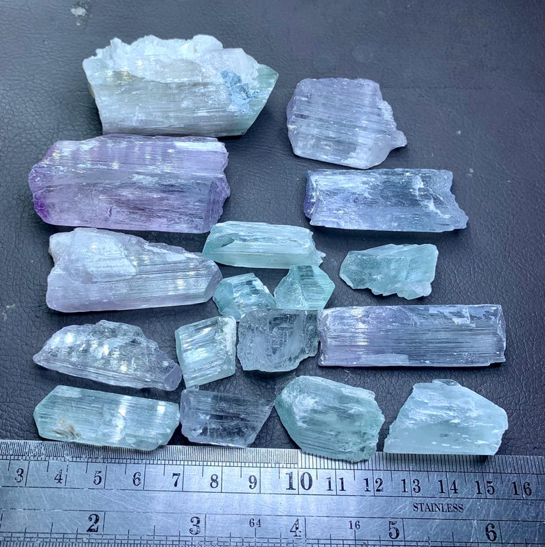 May include: A collection of raw, uncut gemstones in shades of blue, green, and purple. The stones vary in size and shape, with visible crystalline structures. A metal ruler with inch markings is at the bottom of the image.