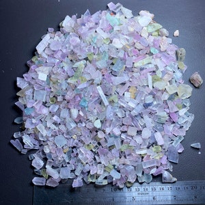 Puede incluir: Un montón grande de pequeños cristales translúcidos multicolores. Los cristales son principalmente transparentes, con algunos matices de rosa, azul y verde. Los cristales están dispersos sobre una superficie gris oscuro.