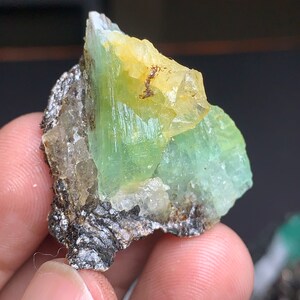 Op de afbeelding: Een groen en geel kristalspecimen met een ruwe, natuurlijke textuur. Het kristal is bevestigd aan een donkergrijze rots.