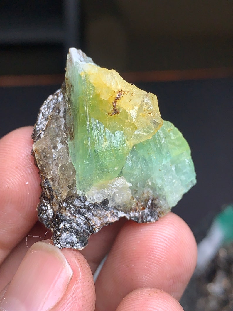 Op de afbeelding: Een groen en geel kristalspecimen met een ruwe, natuurlijke textuur. Het kristal is bevestigd aan een donkergrijze rots.