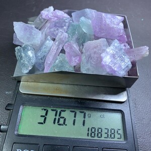 以下が含まれることがあります： デジタルスケールには376.77グラムと1883.85カラットが表示されています。金属製のトレイには、ラベンダー、ブルー、グリーンの色合いの、生の半透明の結晶が集められています。スケールには、PCS、ON/OFF、MODE、TAREとラベル付けされたボタンがあります。