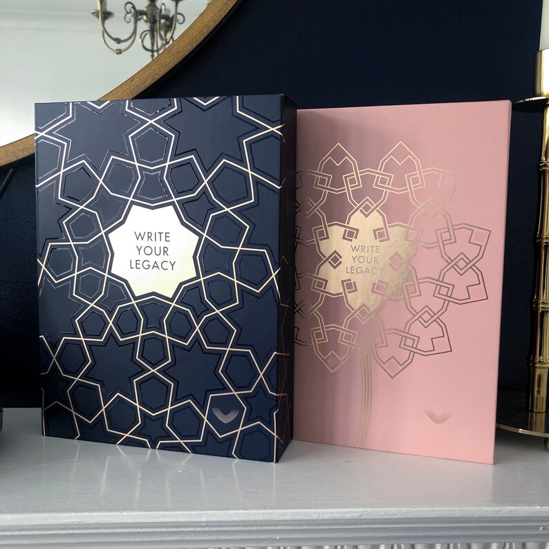 May include: Two hardcover journals with gold foil designs. The journal on the left is navy blue with a gold geometric pattern and the text "Write Your Legacy". The journal on the right is pink with a gold geometric pattern and the text "Write Your Legacy".