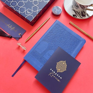 May include: A blue notebook with a gold embossed design of a tree. The notebook is open to a page with the text "Ramadan Kareem" in gold lettering. The notebook is on a red surface with a gold pen, a blue and gold bookmark, and a small gold bookmark with the word "Alhamdulillah" on it.