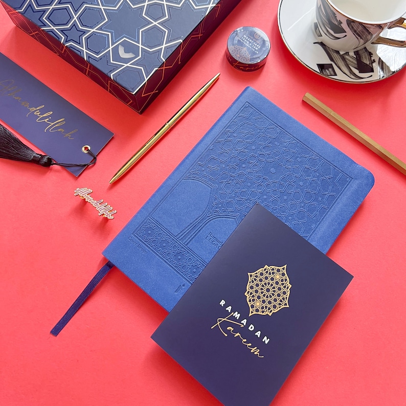 May include: A blue notebook with a gold embossed design of a tree. The notebook is open to a page with the text "Ramadan Kareem" in gold lettering. The notebook is on a red surface with a gold pen, a blue and gold bookmark, and a small gold bookmark with the word "Alhamdulillah" on it.
