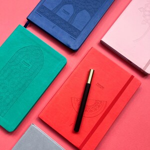 May include: Five different colored notebooks with embossed designs and the words "Remarkable Legacy" on the front. The notebooks are arranged on a pink surface. A black pen is lying on top of one of the notebooks.