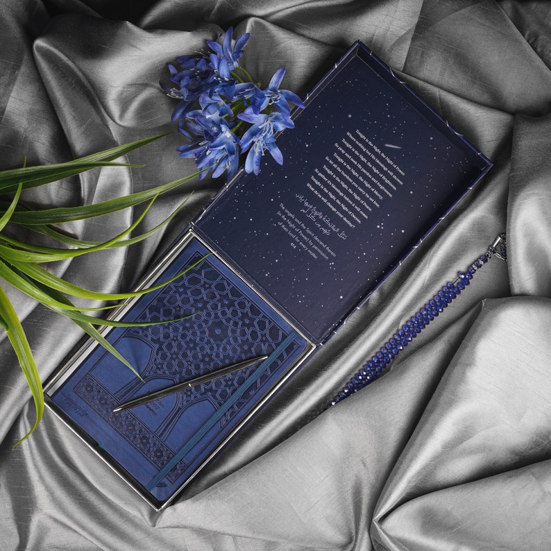 May include: A blue and silver gift box with a blue notebook, pen, and a blue beaded bracelet. The notebook has a star-filled cover with a quote in Arabic script. The box is open and the notebook is visible.