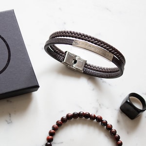 May include: A brown leather bracelet with a silver metal plate engraved with the words "Alhamdulillah". The bracelet is on a white marble surface with a black ring and a red beaded bracelet.