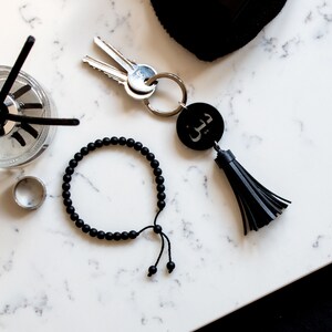 May include: A black beaded bracelet with a tassel keychain featuring an anchor charm. The keychain is attached to a silver key ring.