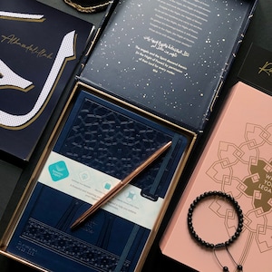 May include: A blue journal with gold accents and a gold pen. The journal has an intricate geometric pattern on the cover. The journal is open to a page with a quote about the Night of Power in Arabic and English. The journal is surrounded by other items, including a pink book with a geometric pattern, a black bracelet, and a black and gold book with the word "Alhamdulillah" written on the cover.
