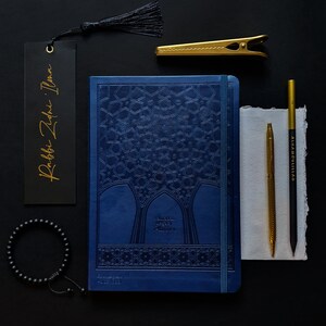 May include: A blue planner with a gold embossed design and the text "Ramadan Legacy Planner" on the cover. The planner is surrounded by various stationery items, including a gold bookmark, a gold pen, a black pencil, and a gold paperclip.