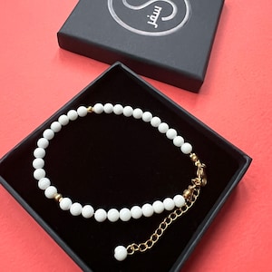 Peut inclure: Bracelet de perles blanches avec des détails dorés, présenté dans un coffret noir. Le bracelet est composé de petites perles blanches rondes et d'éléments métalliques dorés. Le coffret porte un logo argenté et le mot arabe "سفر".