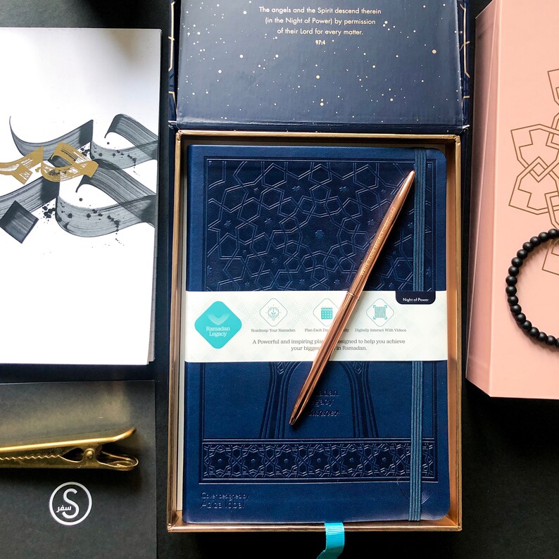 May include: A blue journal with a gold pen and a decorative pattern. The journal is in a box with a gold trim. The box is open and the journal is visible. The journal has the words "Ramadan Legacy" and "Night of Power" printed on it.