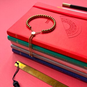 May include: A stack of four red, green, blue, and gray journals with gold lettering that reads "Ramadan Legacy Planner". A gold beaded bracelet with a gold tassel is on top of the journals.