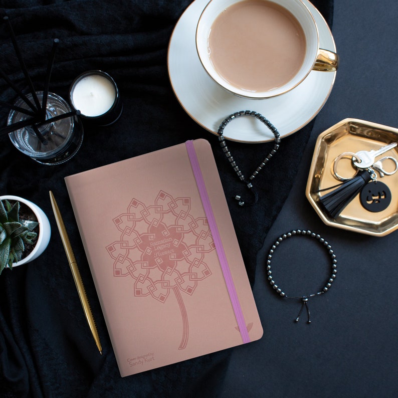 May include: A pink leather journal with a pink elastic closure and a pink and white geometric design. The journal has the text "Ramadan Legacy Journal" on the cover. The journal is on a black surface with a cup of tea, a gold tray with keys, and a black bracelet.