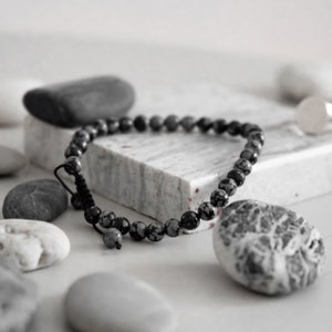 Peut inclure: Un bracelet en perles de pierre tachetées de noir et de blanc avec une fermeture en cordon noir. Le bracelet est posé sur une surface en marbre blanc avec d'autres pierres.