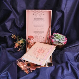 May include: A pink and gold journal with a decorative design on the cover. The journal is open and a white beaded necklace is resting on the pages. There is a small glass jar filled with gold glitter and a copper pen next to the journal. The background is a dark blue fabric.