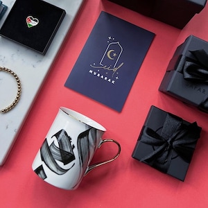 May include: A white ceramic mug with black brushstroke calligraphy and a gold handle, a dark blue card with gold Eid Mubarak text, a heart-shaped pin with the Jordanian flag, a gold beaded bracelet, and two black gift boxes with black ribbons.