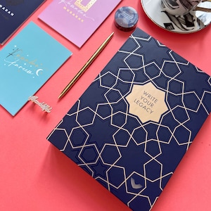 May include: A navy blue journal with a gold geometric pattern and the text "Write Your Legacy". The journal is on a pink surface.