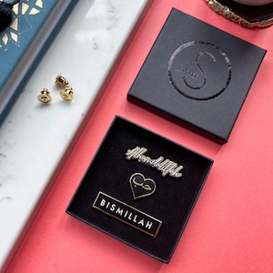 May include: Two gold-colored enamel pins in a black box. The pins feature the Arabic words "Alhamdulillah" and "Bismillah" in white lettering. The box is lined with black velvet and has a gold-colored heart-shaped pin with the Arabic word for "love" in the center.