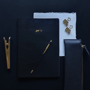 May include: A black leather journal with gold lettering that says "دارو" on the front cover. A gold pen and a gold paper clip are also in the image.